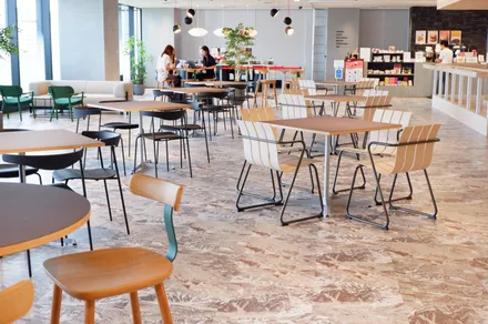 モダンなカフェテリアまたはオフィスの共有スペース。木製のテーブルと椅子、大理石の床、緑の植物、背景に本棚がある。数人の人々が奥のテーブルで作業している。