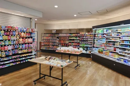 コンビニエンスストアの内部。多くの棚に様々な食品や飲料が並んでいる。手前には商品を展示するテーブルがある。床は木製で、天井には照明が設置されている。