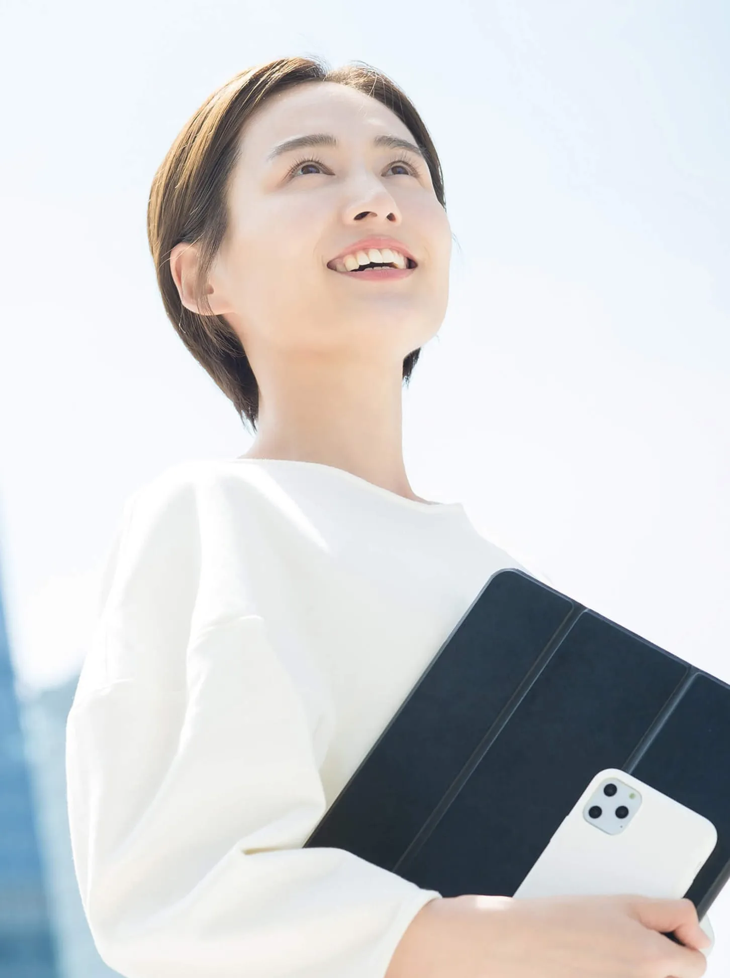 青空を背景に微笑むショートヘアの女性、白いトップスを着用し、黒いタブレットとスマートフォンを持っている