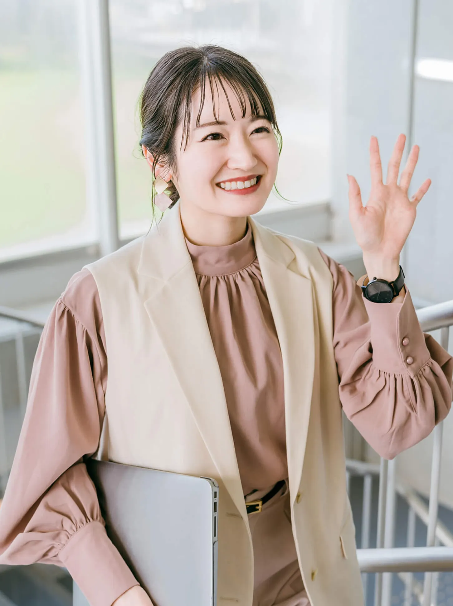 室内で手を振りながら笑顔を見せる女性、ブラウンのブラウスとベージュのベストを着用し、ノートパソコンを持っている