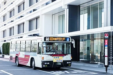 白い建物の前に停車している赤と白の市バス。建物には多くの窓があり、バスの前にはバス停の標識が見える。