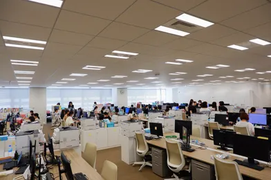 広々としたオフィス空間。天井には多数の蛍光灯が並び、大勢の社員がデスクに向かって作業している。机上にはコンピューターやプリンターが並んでいる。明るく開放的な雰囲気の職場環境。