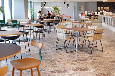 モダンなオフィスの共用スペース。木製のテーブルと椅子、大理石の床、緑の植物、照明器具が配置されている。奥には人々が座っており、販売コーナーも見える。