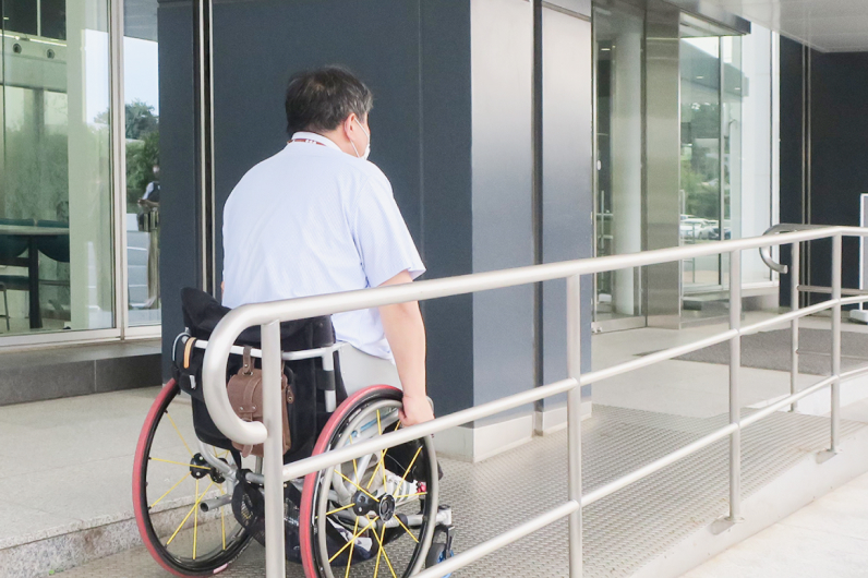 車椅子に乗った男性が、スロープを上がって建物の入り口に向かっている様子。背中側から撮影されており、男性は水色のシャツを着てマスクを着用している。