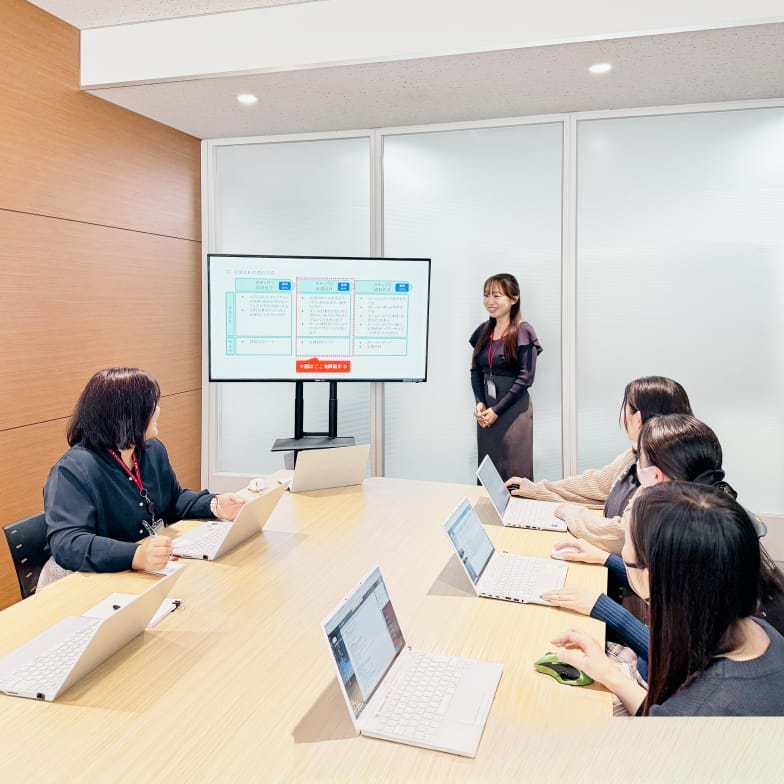 会議室で、女性のプレゼンターが画面に表示された資料について聴衆に説明している様子。複数の参加者がノートPCを使用している。