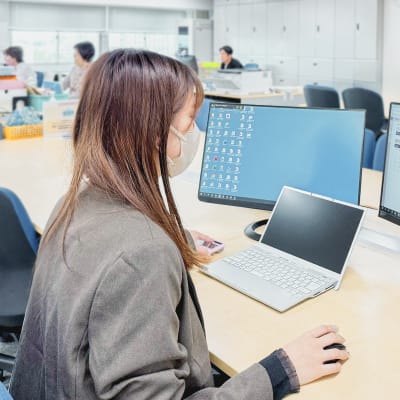オフィスで、マスクを着用した女性がノートPCと外部モニターを使って作業している様子。