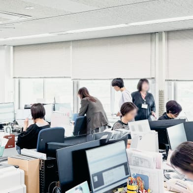 開放的なオフィス内で、複数の従業員が各自のデスクで作業したり、立ち上がって同僚と話したりしている様子。