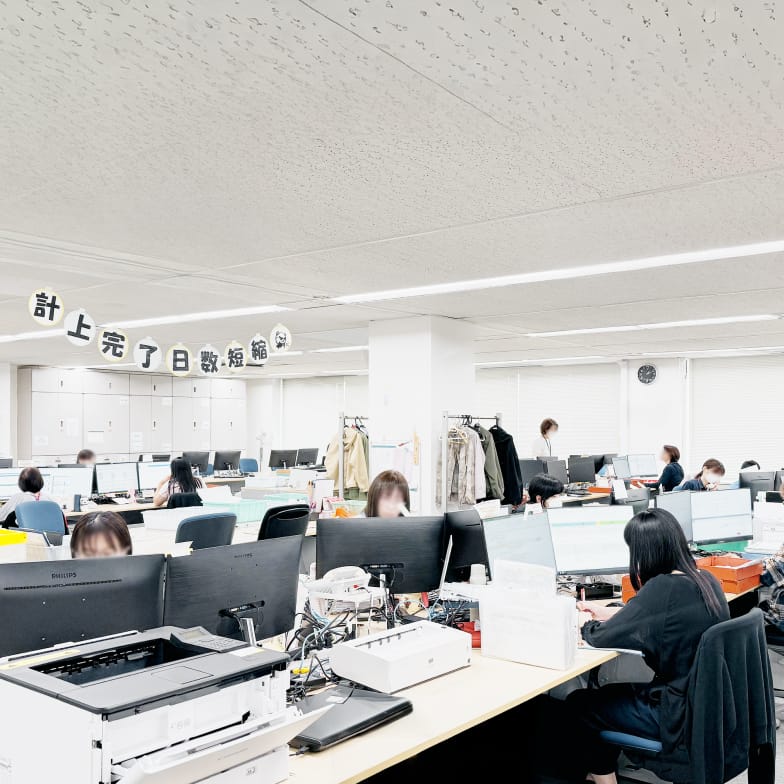 大規模なオフィスフロアで、多くの人々がデスクに向かってPC作業をしている様子。