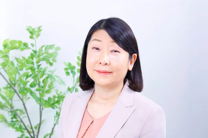 明るいピンクのブラウスの上に薄いピンクベージュのジャケットを着用し、微笑んでいる女性の上半身のポートレート。背景はぼかされており、左側に緑の観葉植物の葉が見える。